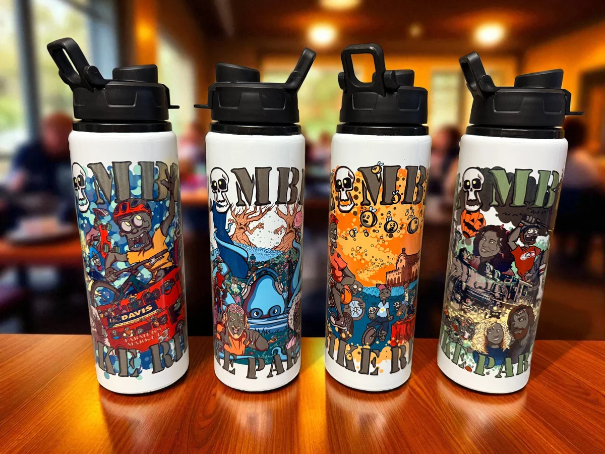 Four white Zombie Bike Parade water bottles on a wooden table, each printed with a different year’s festival artwork.