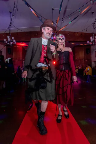 A man in a kilt and a woman in sugar skull makeup walk arm in arm down the red carpet.
