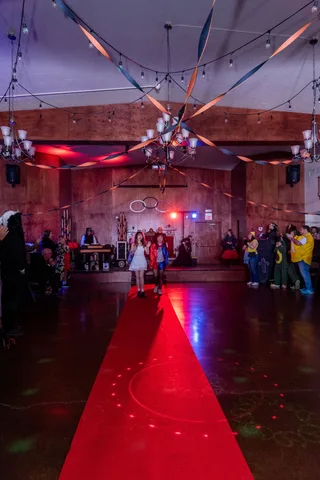 Two costumed teens walk down a lit red carpet at the Zombie Ball as onlookers cheer.