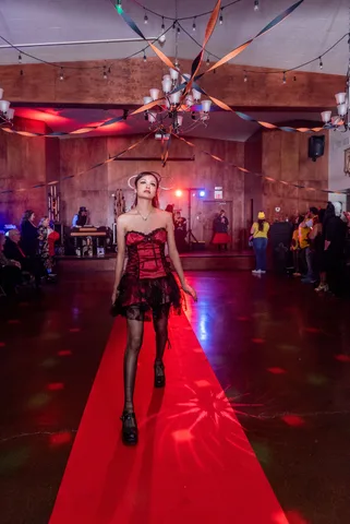 A woman in a red and black corset dress stands sharply lit on the ballroom floor.