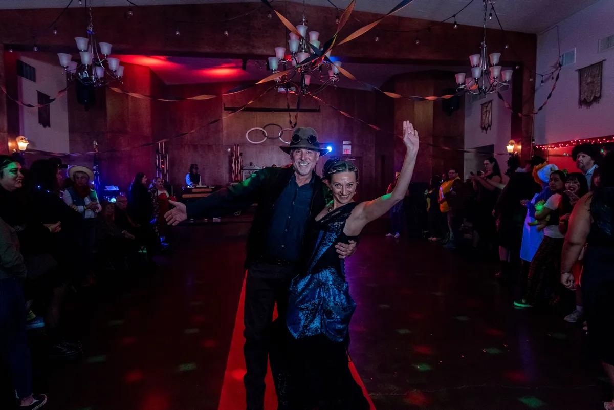 A couple in costume dance on the red carpet, one arm raised, as the crowd looks on.