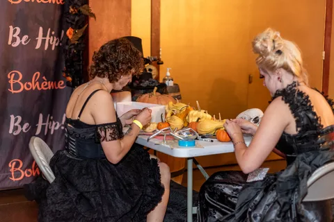 Two guests in gothic gowns decorate small pumpkins and gourds at a table beside the Boheme backdrop.