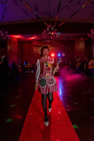 A woman in a picnic basket dress walks the red carpet mid-stride, smiling.