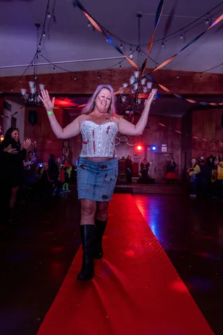 A woman in a bloodied corset and torn denim skirt raises her arms on the red carpet as the crowd cheers.
