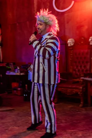 The emcee in a striped suit and wild wig performs under red and blue stage lights.