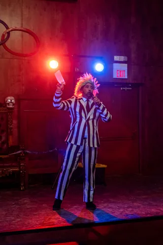 The emcee in a striped suit and wild wig speaks into a microphone under dramatic red and blue lights.