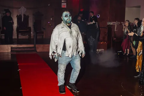 A guest in a torn white zombie costume walks the runway with arms limp.