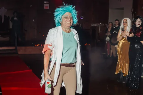 A woman in a teal-haired mad scientist costume smiles while walking the runway.