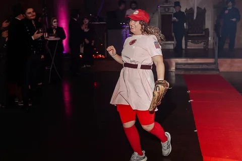 A woman in a softball uniform costume takes a dynamic step mid-stride on the runway.
