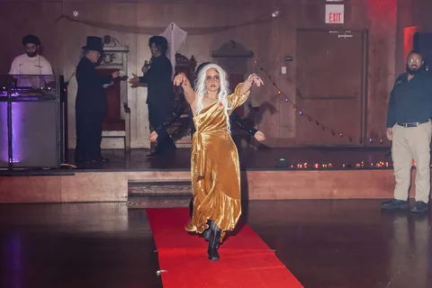A woman in a gold gown with ghostly makeup raises an arm mid-motion on the runway.