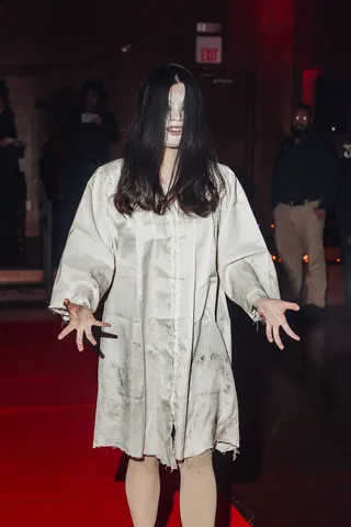 A woman dressed as a long-haired horror ghost reaches toward the camera, hair covering her face.