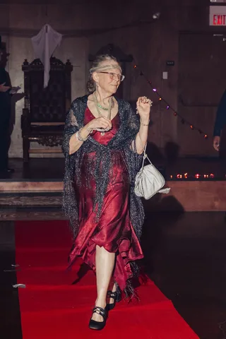 An older woman in a red dress and glasses walks the runway holding her purse.