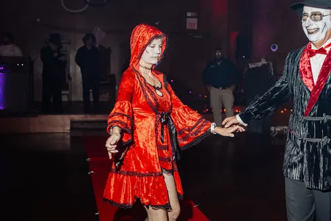 A couple in a red kimono robe and black tuxedo with a white mask hold hands on the runway.