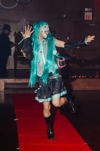 A woman in a teal-haired cosplay costume dances with both arms raised, hair flying.