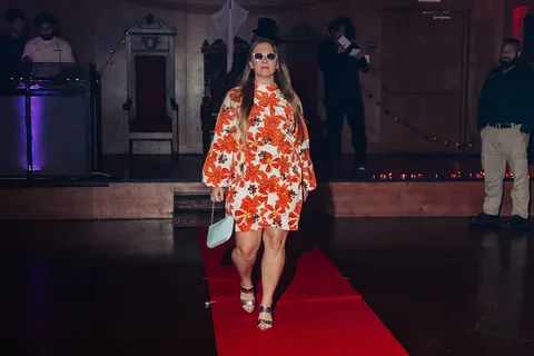 A woman in an orange floral dress smiles while walking closer to the camera.