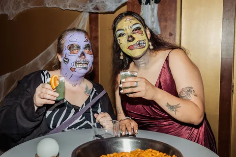 Two women in vivid zombie makeup and gowns share drinks and laughter.