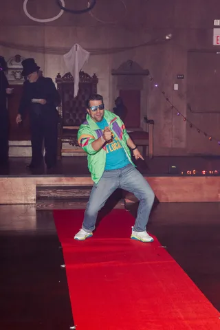 A man in a colorful retro jacket and sunglasses strikes a fun dance pose on the runway.