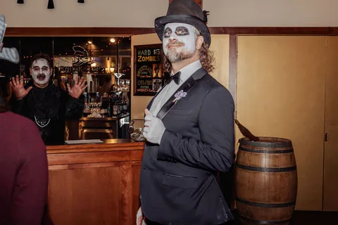 A man in a tuxedo with white face paint waves to the camera from the bar.