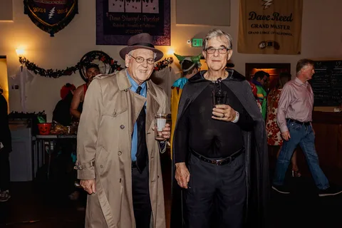 Two older men in trench coats and capes stand together holding drinks at the bar.