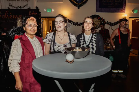 Three women in costume smile together at a small round table.