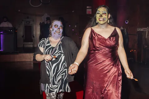 Two women in gowns and makeup hold hands while walking the runway.