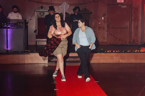 Two women continue down the red carpet runway, distant from camera.