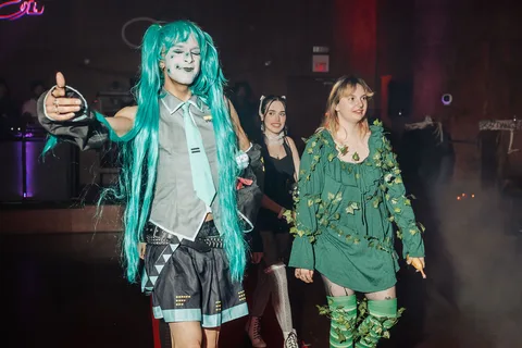 A woman in a teal wig gives a thumbs up on the red carpet runway with friends behind her.
