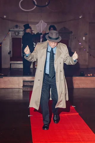 An older man in a trench coat gives a thumbs up while walking the runway.