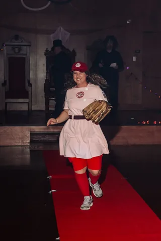 A woman in a softball uniform costume smiles broadly while walking the runway.