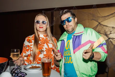 A couple in colorful retro costumes and sunglasses smile while holding drinks and food.
