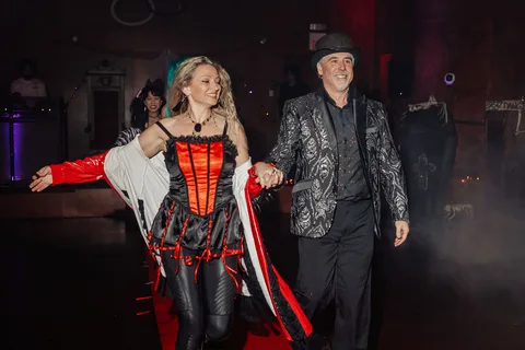 A woman in a red and black corset dances with a man in a gold jacket, arms outstretched.