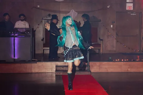 The teal-haired cosplay dancer leaps mid-air on the runway.