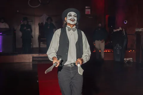 A man with joker-style face paint holds a prop knife while standing on the red carpet.