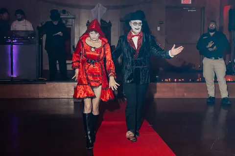The costumed couple walks with arms extended along the runway.