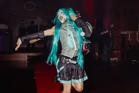 A woman in a teal-haired cosplay costume raises an arm mid-dance on the runway.