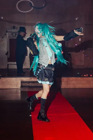 The teal-haired cosplay dancer spins mid-motion on the runway.