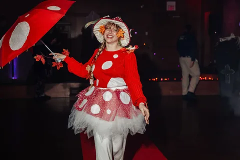 A woman in a mushroom costume smiles brightly while holding an umbrella on the runway.