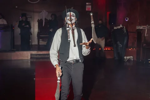 A man with joker-style face paint performs mid-step with a cane on the runway.