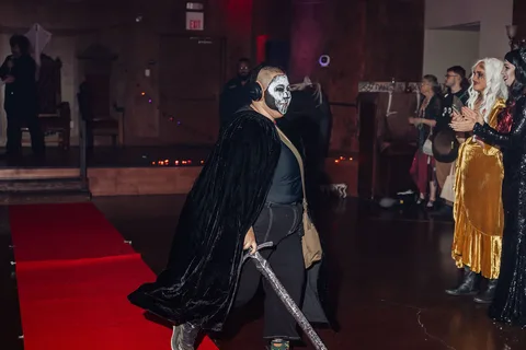 A figure in a dark hooded robe and skull mask poses with a cane on the runway.
