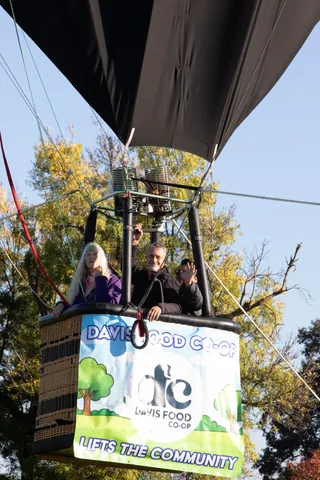 Balloon riders look down over the basket rail above a Davis Food Co-op banner.