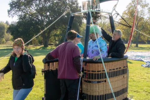 Two riders stand in the balloon basket beside a partial Lifts the Community banner.