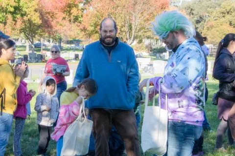 A costumed volunteer with teal hair hands a bag to a bald man as a boy waits behind.