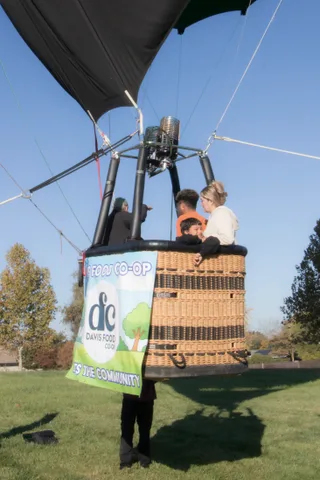 Three riders in the basket drift over the treetops beneath the Davis Food Co-op banner.