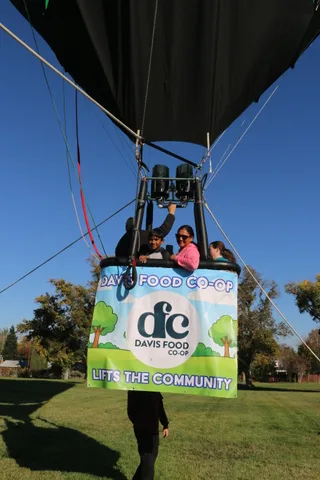 A small group including a child rides in the balloon basket above the sponsor banner.
