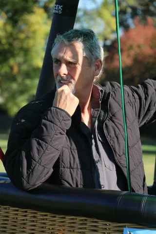A gray-haired man rests his hand on his chin in a thoughtful outdoor portrait.