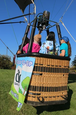 A rider in a pink jacket is mostly hidden behind a sponsor banner on the balloon basket.