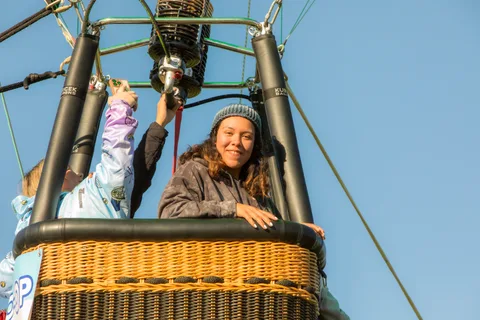 A woman in a beanie smiles and waves from the basket beside a child wrapped in a blanket.