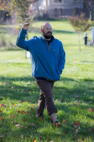 A bald man in a blue fleece raises his arm mid-gesture in the cemetery park.