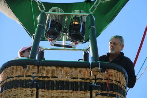 Two men stand in the balloon basket beneath the burners against blue sky.