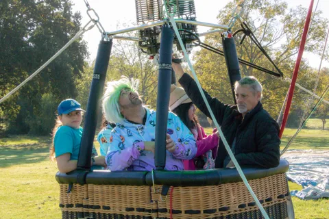 A man in a green wig leans back playfully in the balloon basket as others look on.